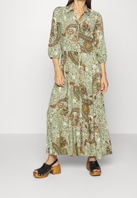 Abito lungo verde con un motivo paisley in tonalità arancioni e marroni, con chiusura frontale a bottoni e maniche puff a 3/4. Zoccoli neri completano il look.