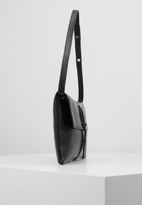 Černá kožená kabelka s texturou na rameno, s nastavitelným popruhem, zavíráním na klopu a stříbrným detailem na sponě, vystavená na bílém podstavci.