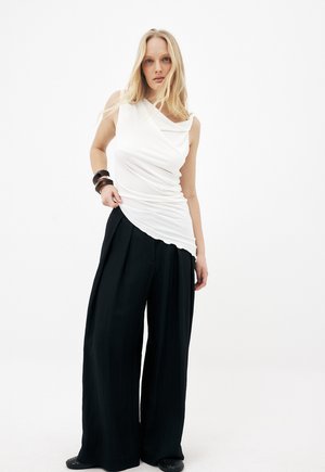 Femme aux longs cheveux blonds portant un haut blanc asymétrique sans manches, un pantalon noir à jambes larges, et des bracelets foncés au poignet gauche.