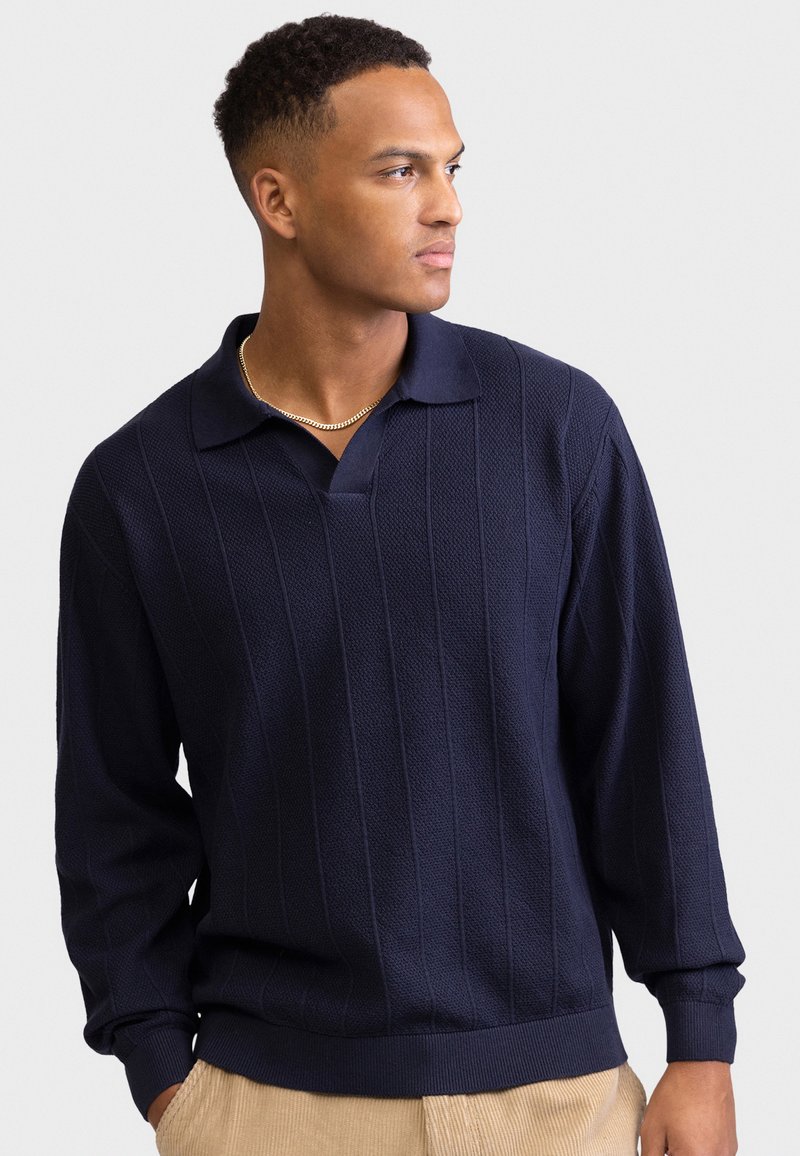 Άνδρας που φοράει μπλε πουλόβερ πόλο με υφή και μακρυμάνικο σε σκούρο μπλε και μπεζ παντελόνι, κοιτάζοντας προς τα πλάγια σε απλό ανοιχτόχρωμο φόντο.