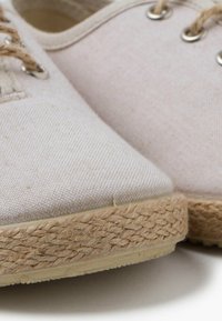 Espadrillas in tela beige chiaro con punte arrotondate, dotate di suole in corda di juta e occhielli metallici per i lacci. Dettaglio in tessuto texture visibile.