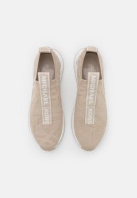 Beżowe dzianinowe trampki z teksturowanym wzorem, wyposażone w białe, markowe elastyczne paski oraz białą, teksturowaną podeszwę.