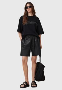 Camiseta negra de gran tamaño con logo texturizado, combinada con pantalones cortos de cuero negros, sandalias negras y un bolso tote negro. Un look suave y casual.