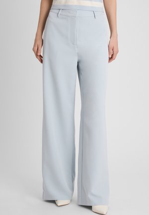 Vrouw draagt lichtblauwe wijde broekspijpen en witte puntige schoenen, staand tegen een effen lichtgrijze achtergrond.