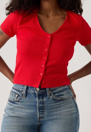 Femme portant un haut rouge côtelé près du corps à manches courtes avec boutons, associé à un jean en denim bleu taille haute, mains sur les hanches.
