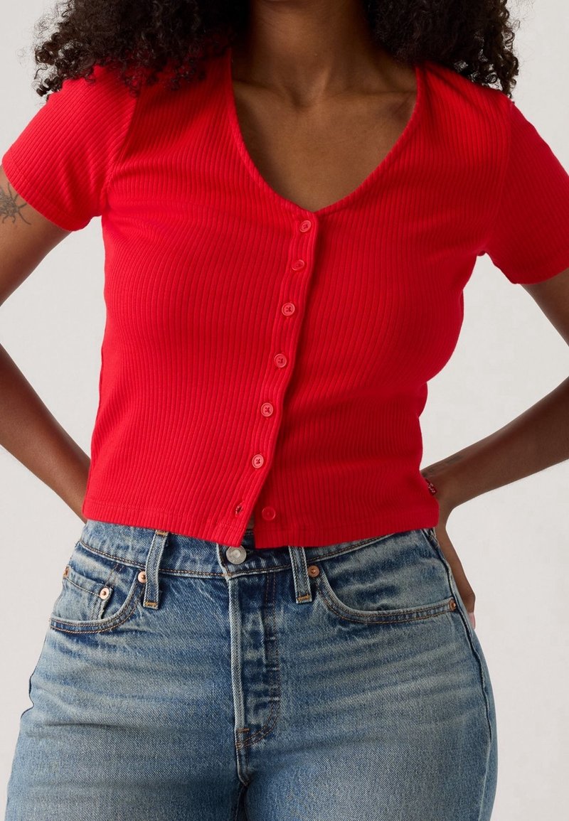 Femme portant un haut rouge côtelé près du corps à manches courtes avec boutons, associé à un jean en denim bleu taille haute, mains sur les hanches.