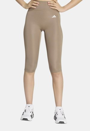 Donna che indossa leggings beige fino al ginocchio con logo Adidas bianco, in piedi con le mani sui fianchi e scarpe da ginnastica Adidas bianche con strisce nere.