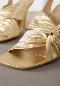 Gouden hoge hakken sandalen met een gedraaid knoopontwerp aan de voorkant, een gladde metallic leren textuur en open teen stijl.