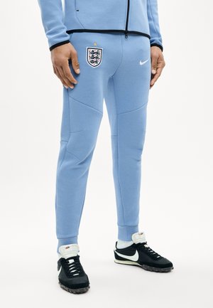 Personne portant un pantalon de survêtement bleu clair de l'équipe d'Angleterre avec logo Nike, chaussures de sport Nike noires et blanches sur fond blanc.