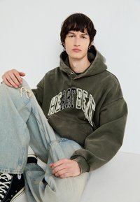 Olajzöld kapucnis pulóver, „HEARTBEAT” grafikával, elhasználódott betűtípussal, világoskék, bő nadrággal és fekete sneaker cipővel párosítva.