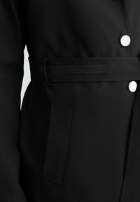 Cappotto nero con una texture liscia, cintura in vita, tasca a toppa e bottoni in metallo argento per la chiusura.