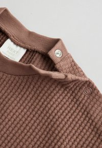 Braun strukturiertes Babyshirt mit geripptem Ausschnitt, Druckknopfverschluss und gewebtem Stoff. Das Etikett trägt die Aufschrift "NEXT baby."