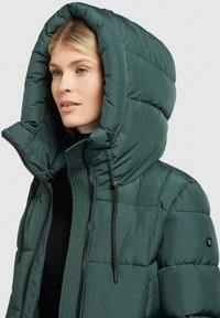 Zals pūkainais mētelis ar lielu kapuci. Iekļauj rāvējslēdzēju priekšpusē, elastīgas auklas un quilted tekstūru. Minimāla zīmola identifikācija uz pleca.