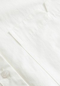 Κοντινή λήψη λευκού υφάσματος πουκάμισου που δείχνει τσέπη στο στήθος, ορατή ραφή και λευκό κουμπί κοντά στη μανσέτα.