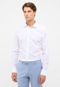 Camicetta bianca con colletto a camicia e maniche lunghe, tessuto liscio e patta dei bottoni nascosta. Abbinata a pantaloni leggeri blu texture.