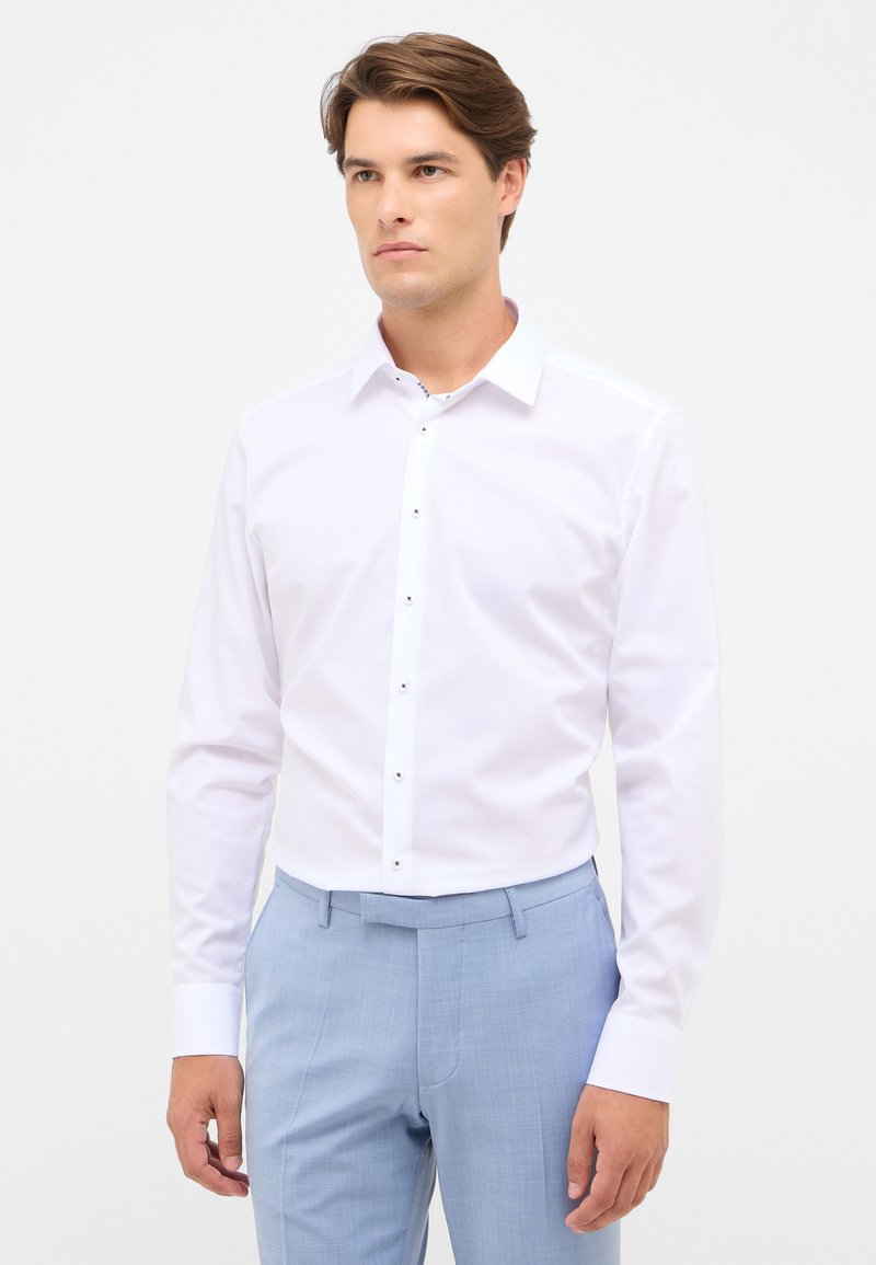 Camicetta bianca con colletto a camicia e maniche lunghe, tessuto liscio e patta dei bottoni nascosta. Abbinata a pantaloni leggeri blu texture.