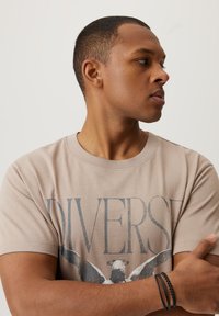 Beżowy bawełniany t-shirt z napisem "DIVERSE" w ciemnoszarym kolorze. Krótkie rękawy, okrągły dekolt, noszony przez osobę z założonymi rękami.