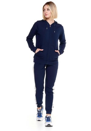 Une femme porte un sweat à capuche bleu marine zippé et un pantalon de jogging assorti à bandes blanches sur les côtés, ainsi que des baskets bleues, sur fond blanc.