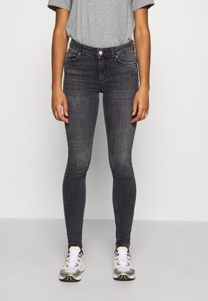 Person iført sorte skinny jeans, grå t-shirt og hvide sneakers står mod en ensfarvet lys baggrund.