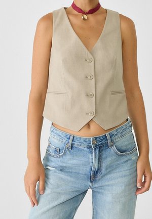 Mouwloos beige gilet met knopen, gedragen met lichtblauwe spijkerbroek en een rode chokerketting met een ronde gouden hanger.
