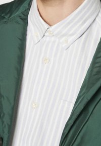 Vihreä takki, jonka alla on valkoinen kauluksellinen paita, jossa on vaaleansiniset raidat. Paidassa on tasku ja edessä nappikiinnitys, jossa on kermaisia nappeja.