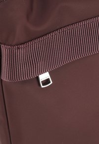 Sac en nylon bordeaux avec un tissu texturé, une large sangle côtelée et une étiquette logo en métal argenté sur le devant.