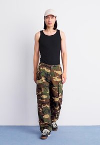 Joven con una camiseta sin mangas negra, pantalones cargo de camuflaje, gorra beige y zapatillas negras caminando hacia adelante frente a un fondo blanco liso.