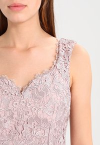 Lichtroze jurk met een bloemenkant overlay, gegolfde halslijn en delicate geborduurde details langs de bandjes en het lijfje.