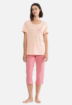 Žena s tmavými vlasy po ramena, oblečená v světle růžovém tričku s krátkými rukávy a potiskem palem a středně dlouhých růžových kalhotách, bosá.