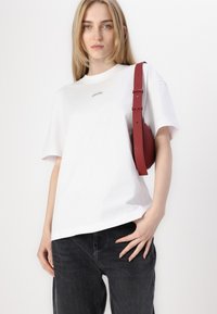 T-shirt en coton blanc avec un col rond et des manches courtes, arborant un petit logo. Associé à un jean gris foncé et un sac rouge.