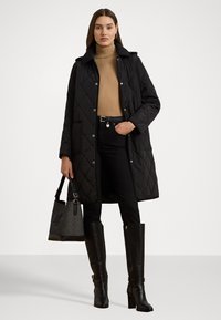 Zwarte gewatteerde jas met een kraag, lange mouwen en zakken. Gekozen boven een beige turtleneck en gecombineerd met zwarte jeans en hoge zwarte laarzen.