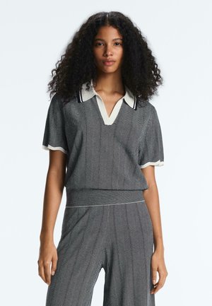 Woman with curly hair wearing a grey patterned short-sleeve top with white collar and matching wide-leg pants against a plain background.