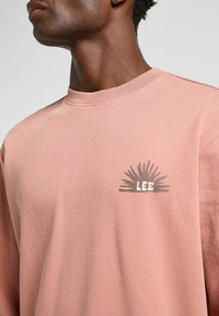 Różowa bawełniana bluza z okrągłym dekoltem. Wyraźny nadruk graficzny "LEE" z promienistym wzorem w brązie na lewej stronie klatki piersiowej.