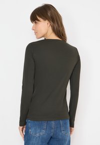 Dunkelgrünes Langarmhemd aus glattem Stoff, mit rundem Ausschnitt und taillierter Silhouette, kombiniert mit hellblauen Jeans.