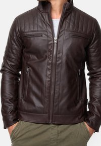 Brun læderjakke med quiltede skuldre, to forlommer med lynlås og høj krave. Har en glat tekstur og et tætsiddende design.