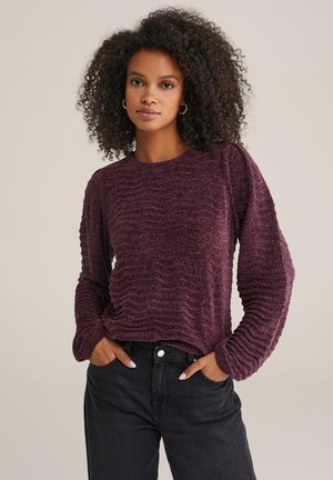 Maglione bordeaux con motivo texturizzato e ondulato e maniche lunghe, abbinato a jeans neri. Design semplice con scollatura a girocollo.