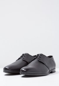 Chaussures en cuir noir à texture lisse, bouts arrondis, lignes épurées et trois œillets. Elles présentent un talon bas et un design minimaliste.