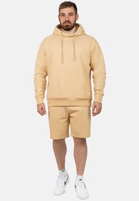 Felpa beige in misto cotone con lacci regolabili, tasca a marsupio frontale e pantaloni corti beige abbinati. Sneakers bianche completano l'outfit.