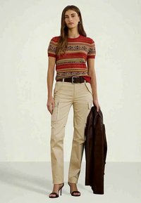 Κόκκινο και μπεζ ριγέ πλεκτό μπλουζάκι, μπεζ cargo παντελόνι με τσέπες, καφέ ζώνη και ένα σκούρο καφέ δερμάτινο μπουφάν κρεμασμένο στον βραχίονα. Παπούτσια με τακούνι.