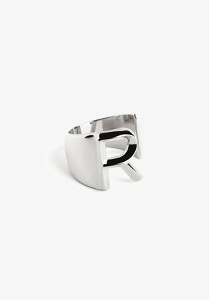 Une bague en métal argenté présente un design de lettre 'R' découpé, une surface lisse et un large bandeau avec une forme moderne et minimaliste.