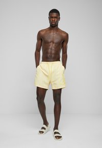 Shorts de bain rayés jaunes avec une taille élastique et des poches, associés à des sandales à enfiler de couleur claire. Texture de tissu lisse.