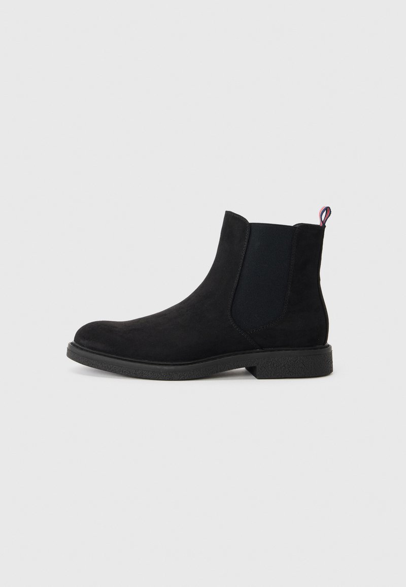 Botín Chelsea negro de ante con paneles laterales elásticos, puntera redonda, suela de goma texturizada y un pequeño tirador de detalle en rojo, blanco y azul.