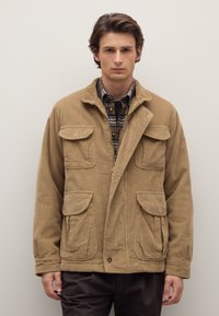 Giacca in velluto a coste color khaki con chiusura a zip, quattro tasche frontali e collo con rever. Indossata sopra una camicia a scacchi e pantaloni scuri.