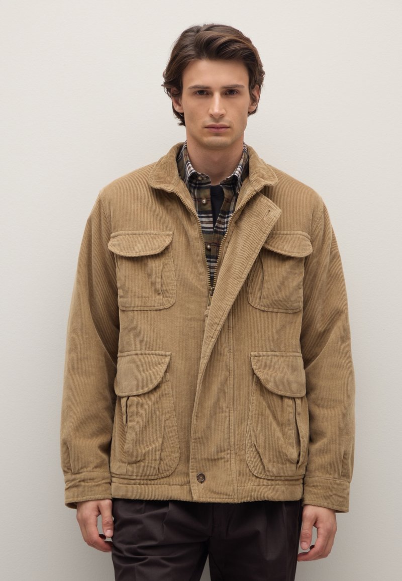 Giacca in velluto a coste color khaki con chiusura a zip, quattro tasche frontali e collo con rever. Indossata sopra una camicia a scacchi e pantaloni scuri.