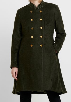 Femme portant un manteau croisé vert foncé avec des boutons dorés, un col montant et des poches fendues, debout avec une main dans la poche.
