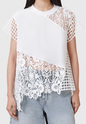 T-shirt blanc avec une superposition en dentelle texturée présentant des motifs floraux et des découpes géométriques, manches courtes et encolure ronde.