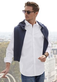 White button-up shirt with a navy sweater draped over the shoulders. Blue jeans, dark sunglasses, and a watch visible on the wrist.