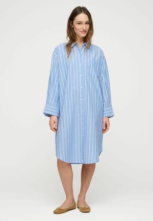 Žena stojící v volných modro-bílých šatech s vertikálními pruhy na knoflíky a béžových balerínách na jednobarevném pozadí.