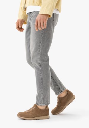 Mężczyzna w jasnoszarych dżinsach, beżowej kurtce i brązowych zamszowych trampkach stojący na białym tle.