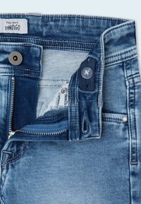 Jeans de mezclilla azul con un diseño desgastado, que cuentan con una cremallera y cierre de botón. En el interior, se puede ver un forro de bolsillo blanco con un patrón.